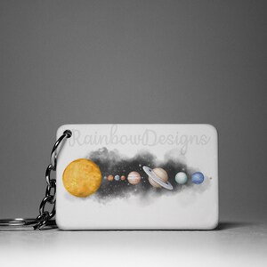 May include: A white rectangular keychain featuring a watercolour illustration of the solar system. The sun is yellow, the planets are various colours, and the background is black with white stars. The text "Rainbow Designs" is printed on the keychain.