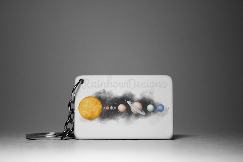 May include: A white rectangular keychain featuring a watercolour illustration of the solar system. The sun is yellow, the planets are various colours, and the background is black with white stars. The text "Rainbow Designs" is printed on the keychain.