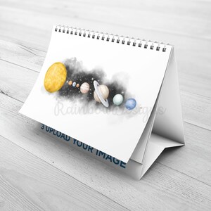 May include: A white desktop calendar featuring a watercolour illustration of the solar system. The sun is yellow, the planets are in various colours, and there is a black and white nebula in the background. The text "3 UPLOAD YOUR IMAGE" is printed on the bottom of the calendar.