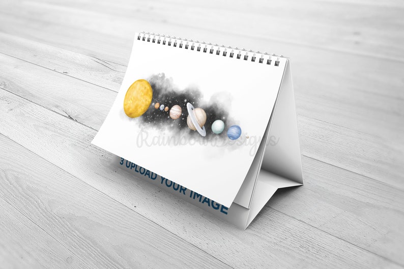 May include: A white desktop calendar featuring a watercolour illustration of the solar system. The sun is yellow, the planets are in various colours, and there is a black and white nebula in the background. The text "3 UPLOAD YOUR IMAGE" is printed on the bottom of the calendar.