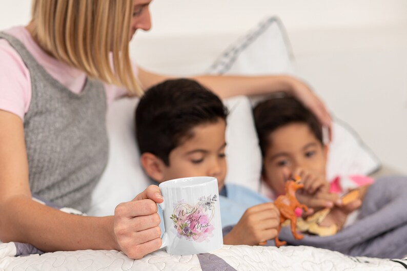 May include: A white ceramic mug with a floral design and the text "World's Best Mom" in a pink heart. The mug is being held by a person's hand. Two children are sitting in the background.