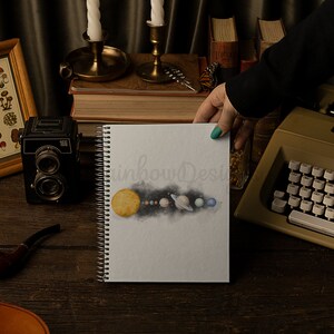 May include: A white spiral bound notebook featuring a watercolour illustration of the solar system. The illustration depicts the sun, planets, and a dark blue nebula. The notebook is on a wooden table with other items, including a vintage camera, a pipe, and a typewriter.