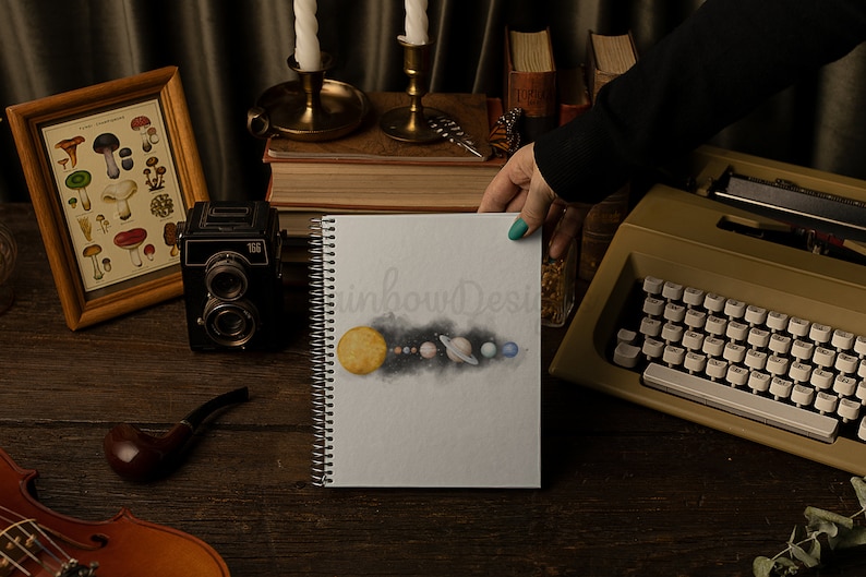 May include: A white spiral bound notebook featuring a watercolour illustration of the solar system. The illustration depicts the sun, planets, and a dark blue nebula. The notebook is on a wooden table with other items, including a vintage camera, a pipe, and a typewriter.