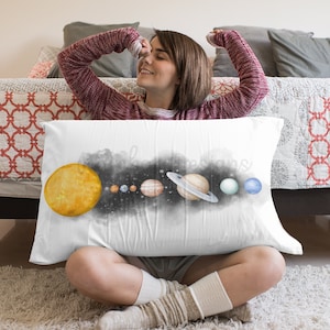 May include: A white pillow featuring a watercolour illustration of the solar system. The sun is yellow, the planets are various shades of blue, green, and brown, and the background is black with white stars. The pillow is resting on a white rug.
