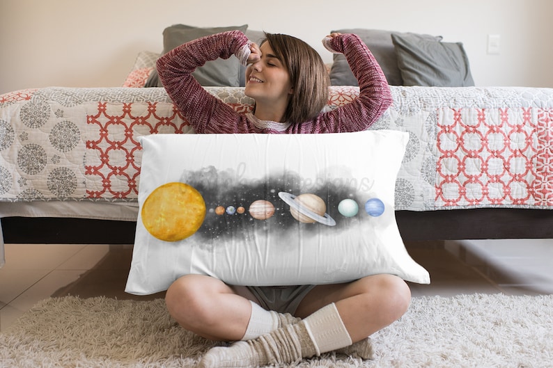 May include: A white pillow featuring a watercolour illustration of the solar system. The sun is yellow, the planets are various shades of blue, green, and brown, and the background is black with white stars. The pillow is resting on a white rug.