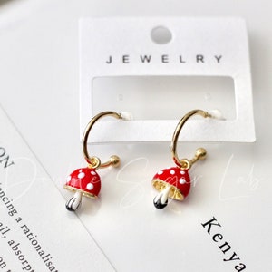 Peut inclure: Une paire de boucles d'oreilles créoles dorées avec des breloques en forme de champignons rouges et blancs. Les boucles d'oreilles sont sur une carte blanche avec le texte "JEWELRY", "WHITE", et "Kenya Hara".