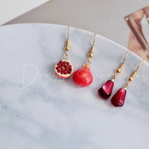 Peut inclure: Trois paires de boucles d'oreilles pendantes avec des crochets dorés. Une paire présente une moitié de grenade rouge, une autre une grenade rouge entière et la dernière paire a deux pierres rouges en forme de larme.