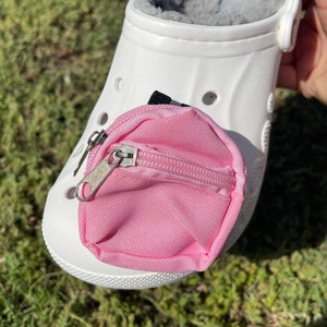 Puede incluir: Una pequeña bolsa rosa con cremallera unida a un zueco blanco. La bolsa está hecha de un tejido suave y tiene una cremallera plateada.