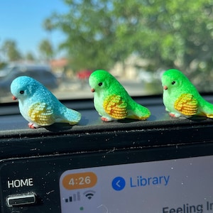 May include: Three small, decorative bird figurines in shades of blue and green with yellow and orange accents. The birds are perched on a dark surface, likely a car dashboard. The background shows a blurred view of a sunny day.