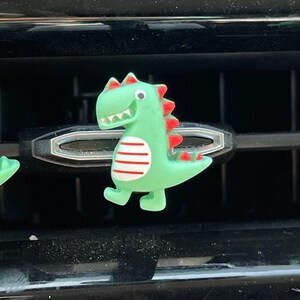 May include: Three green dinosaur-shaped car air vent clips. Each dinosaur has red spines, white and red striped bellies, and a smiling face. The clips are positioned on a black car vent.
