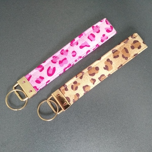 Può includere: Due portachiavi con finiture color oro. Uno è rosa con un motivo leopardato, e l'altro è beige con una stampa leopardata marrone. I portachiavi sono in tessuto e sono progettati per essere indossati al polso.