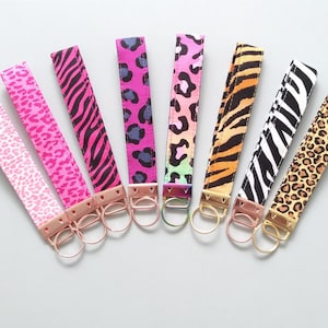 May include: Eight key fobs with animal print designs, including leopard, zebra, and cheetah patterns. The fobs feature pink, black, orange, and white colours. Each has a metal ring and a fabric loop for attaching to keys or bags.