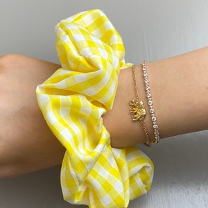 Könnte beinhalten: Ein gelber und weißer Gingham-Haargummi mit einem goldenen Elefanten-Anhänger-Armband an einem Handgelenk.