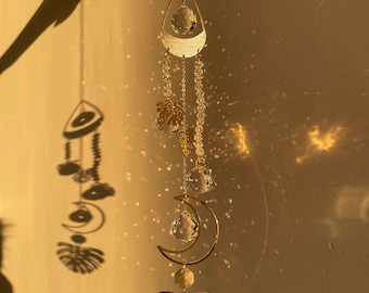May include: A gold crystal suncatcher with a teardrop-shaped crystal, a crescent moon, and a tropical leaf design. The suncatcher is hanging from a chain and casts rainbows on the wall.