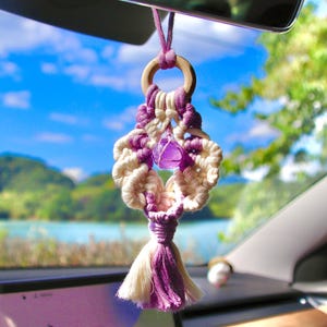 Puede incluir: Un amuleto de coche de macramé morado y blanco con un cristal morado que cuelga de un anillo de madera. El amuleto está colgado del espejo retrovisor de un coche.