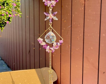 May include: A gold crystal suncatcher with amethyst crystals, a crystal ball, and two crystal teardrops hanging from a gold chain. The suncatcher is hanging from a wooden fence.