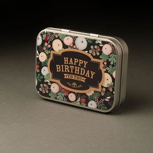 Può includere: Una scatola di metallo argentata con sfondo nero e un motivo floreale. La scatola ha un'etichetta dorata con il testo "Happy Birthday to You".