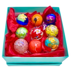 Peut inclure: Un ensemble de neuf billes en verre colorées dans une boîte cadeau turquoise avec du papier de soie rouge. Les billes présentent divers motifs et designs, notamment des volutes, des points et des fleurs.