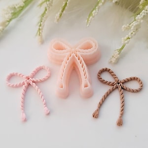 May include: Three decorative bow-shaped items in shades of pink and brown. The bows have a rope-like texture and are arranged on a white surface. The largest bow is a light pink colour, while the other two are a lighter pink and a brown colour.