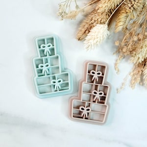 以下が含まれることがあります： プレゼントの積み重ねの形をしたプラスチック製のクッキー型が2つ。左側の型は薄い青色で、右側の型は薄い茶色です。