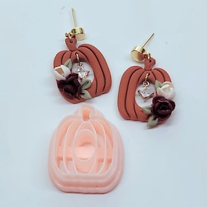 May include: A pair of brown pumpkin-shaped earrings with white and burgundy flowers and a clear rhinestone accent. The earrings are shown with a pink pumpkin-shaped mold.