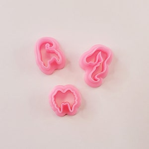 Puede incluir: Tres cortadores de galletas de plástico rosa con forma de fantasma, hongo y lazo.