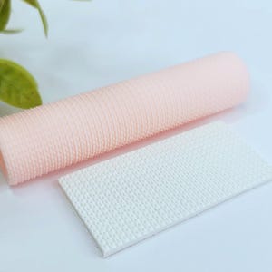 Puede incluir: Un juego de dos herramientas de decoración de pasteles de silicona. Una es un rodillo rosa con una superficie texturizada. La otra es una estera rectangular blanca con una superficie texturizada.