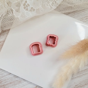 Può includere: Due tagliabiscotti a forma di pane in plastica rosa. I tagliabiscotti sono sovrapposti, con il tagliabiscotti più grande in basso e il più piccolo in alto.