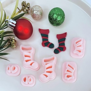 Puede incluir: Un conjunto de ocho cortadores de galletas de plástico rosa con forma de calcetines de Navidad y zapatos de duendes. Los cortadores están sobre una superficie blanca con adornos navideños rojos y verdes.