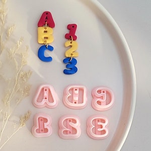 Pode incluir: Um conjunto de cortadores de biscoitos de letras e números rosa. Os cortadores têm a forma das letras A, B e E e os números 1, 2 e 3. Há também dois brincos com as letras A, B e C e os números 1, 2 e 3 em vermelho, amarelo e azul.