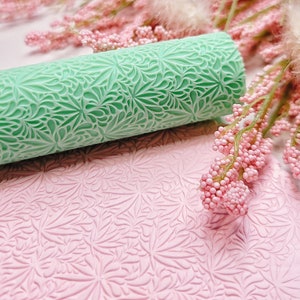 Puede incluir: Un rodillo verde con un patrón floral detallado, descansando sobre una superficie rosa con el mismo patrón. La superficie está estampada con el patrón del rodillo.
