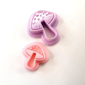 May include: Two mushroom-shaped cookie cutters. One is a light purple colour, and the other is pink with white polka dots. The cutters are made of plastic and are designed for baking or crafting.