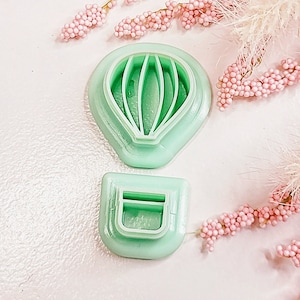 May include: A set of two green plastic cookie cutters shaped like a hot air balloon. The balloon cutter has a detailed design with lines for the basket and the basket cutter has a simple rectangular shape.