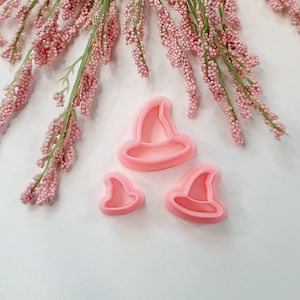 May include: Three pink plastic witch hat cookie cutters of different sizes. The largest cutter is in the center of the image, with a smaller cutter to the left and a smaller cutter to the right.