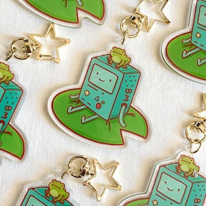 May include: Clear acrylic keychains featuring a teal video game console character with a frog on top, sitting on a green lily pad. Each keychain has a gold star-shaped clasp. The background is a white surface.