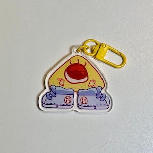 May include: A clear acrylic keychain featuring a cartoon character with a yellow body, red details, and light blue sneakers. The keychain has a yellow clasp for easy attachment. The character has a friendly expression.