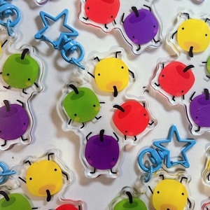 May include: A collection of colorful acrylic keychains featuring cartoon apple designs in red, green, yellow, and purple. Each keychain has a black stem and small black legs. Some keychains have blue star-shaped clasps.