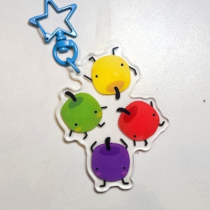 May include: A clear acrylic keychain featuring four cartoon fruit: yellow, green, red, and purple. Each fruit has a black stem, small black feet, and a smiling face. The keychain has a blue star-shaped clasp.