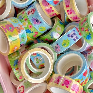 Puede incluir: Una colecci&oacute;n de rollos de cinta washi decorativa en varios colores. La cinta presenta ilustraciones coloridas de animales, frutas y flores. Los rollos son en tonos de azul, amarillo, verde y rosa, perfectos para manualidades y scrapbooking.