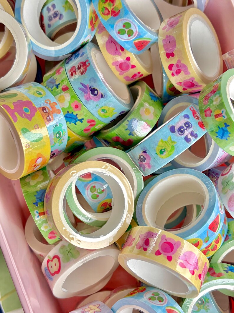 Puede incluir: Una colecci&oacute;n de rollos de cinta washi decorativa en varios colores. La cinta presenta ilustraciones coloridas de animales, frutas y flores. Los rollos son en tonos de azul, amarillo, verde y rosa, perfectos para manualidades y scrapbooking.