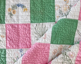 May include: A quilted blanket featuring a checkerboard pattern of solid pink, solid green, and white squares. The white squares are decorated with various embroidered floral and botanical designs in colors including yellow, blue, and green.