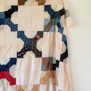 May include: A patchwork quilt featuring a repeating block pattern composed of various fabric squares. The design incorporates a mix of cream-colored background fabrics with small prints and darker, multi-colored patterned patches in blue, red, brown, and black.