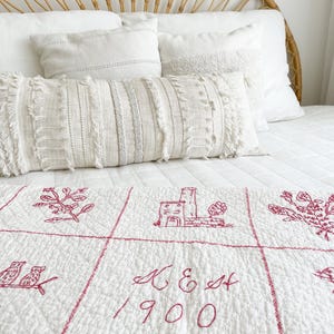 May include: A bed with white bedding, including pillows and a decorative bolster pillow. The bed is topped with a white quilt featuring red embroidered designs of buildings, flowers, and birds, along with the text "K & E Est 1900."