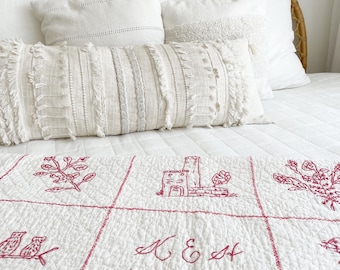 May include: A bed with white bedding, including pillows and a decorative bolster pillow. The bed is topped with a white quilt featuring red embroidered designs of buildings, flowers, and birds, along with the text "K & E Est 1900."