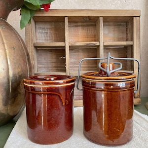 May include: Two brown ceramic crock pots with lids. The crock pot on the right has a metal wire bail handle.