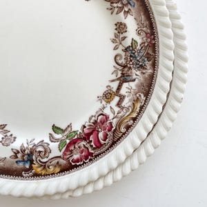 May include: A stack of white ceramic plates with a brown floral border. The plates have a scalloped edge and a detailed design featuring flowers in shades of red, blue, and yellow. The plates are stacked on a white surface.