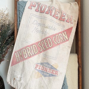 May include: Vintage Pioneer Hybrid Seed Corn sack. The off-white sack features red and blue text, including "PIONEER" and "HYBRID SEED CORN." The sack is draped over a wooden ladder and a blue blanket, creating a rustic, farmhouse aesthetic.