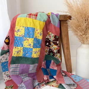 May include: A patchwork quilt featuring a variety of fabric squares in colors including yellow, blue, red, purple, and green. The quilt displays diverse patterns such as floral prints, plaid, and solid colors, with a prominent dusty rose border.