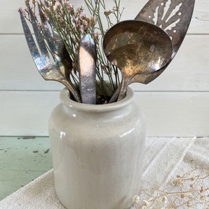 May include: A white ceramic crock with a silver fork, knife, and serving spoon inside. The crock is filled with dried pink flowers.