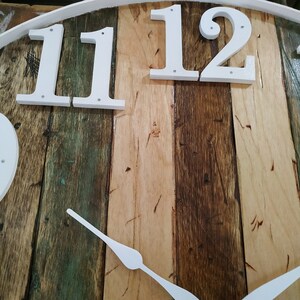 May include: A white clock face with black Roman numeral hour markers on a rustic wooden background. The clock face is made of wood planks in various shades of brown, green, and beige. The clock hands are white and point to the numbers 11 and 12.